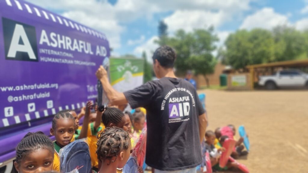 An Ashraful Aid volunteer handing out school bags to children beside the Ashraful Aid van