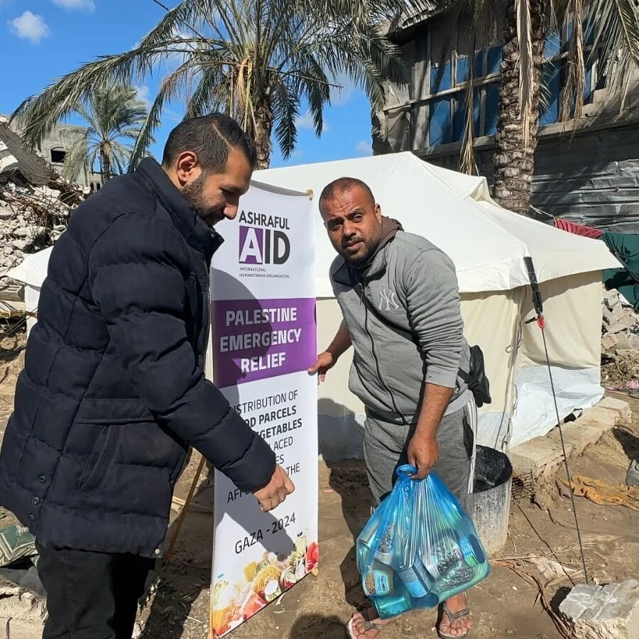 Ashraful Aid team member with a Palestine Emergency Relief banner handing groceries to a man beside a tent in Gaza