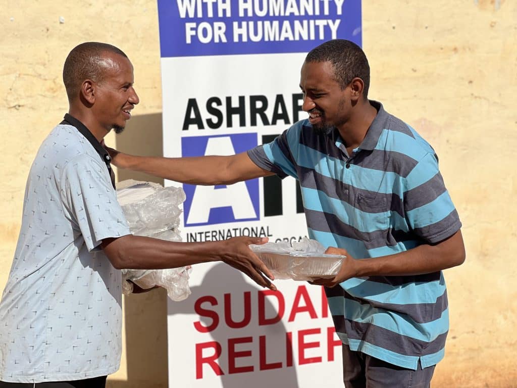 Ashraful Aid volunteer handing a hot meal tray to a man in front of a Sudan Relief banner